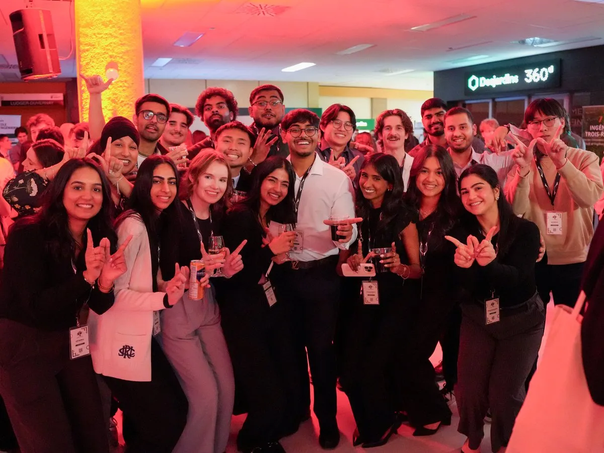 A crowd of students in business attire smiles and makes hand signs at an evening reception.