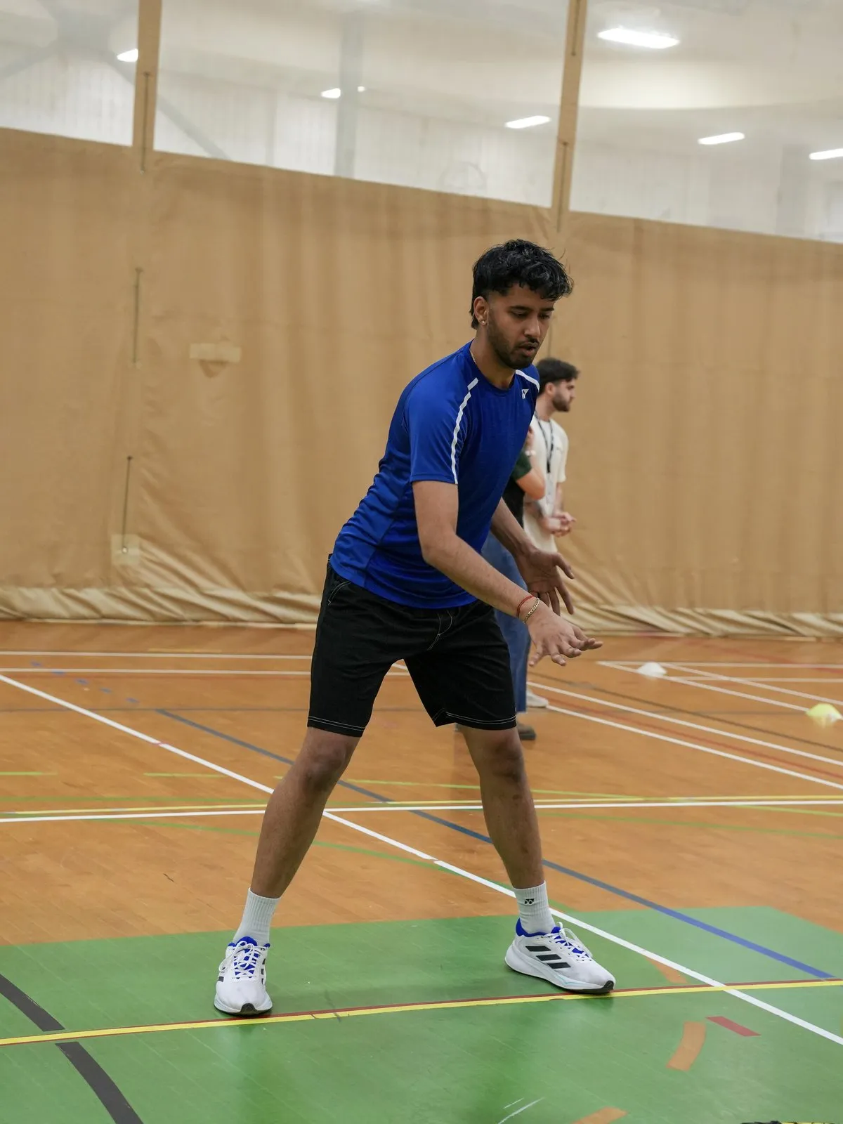 A student in a blue athletic shirt crouches, ready, on a gym court.