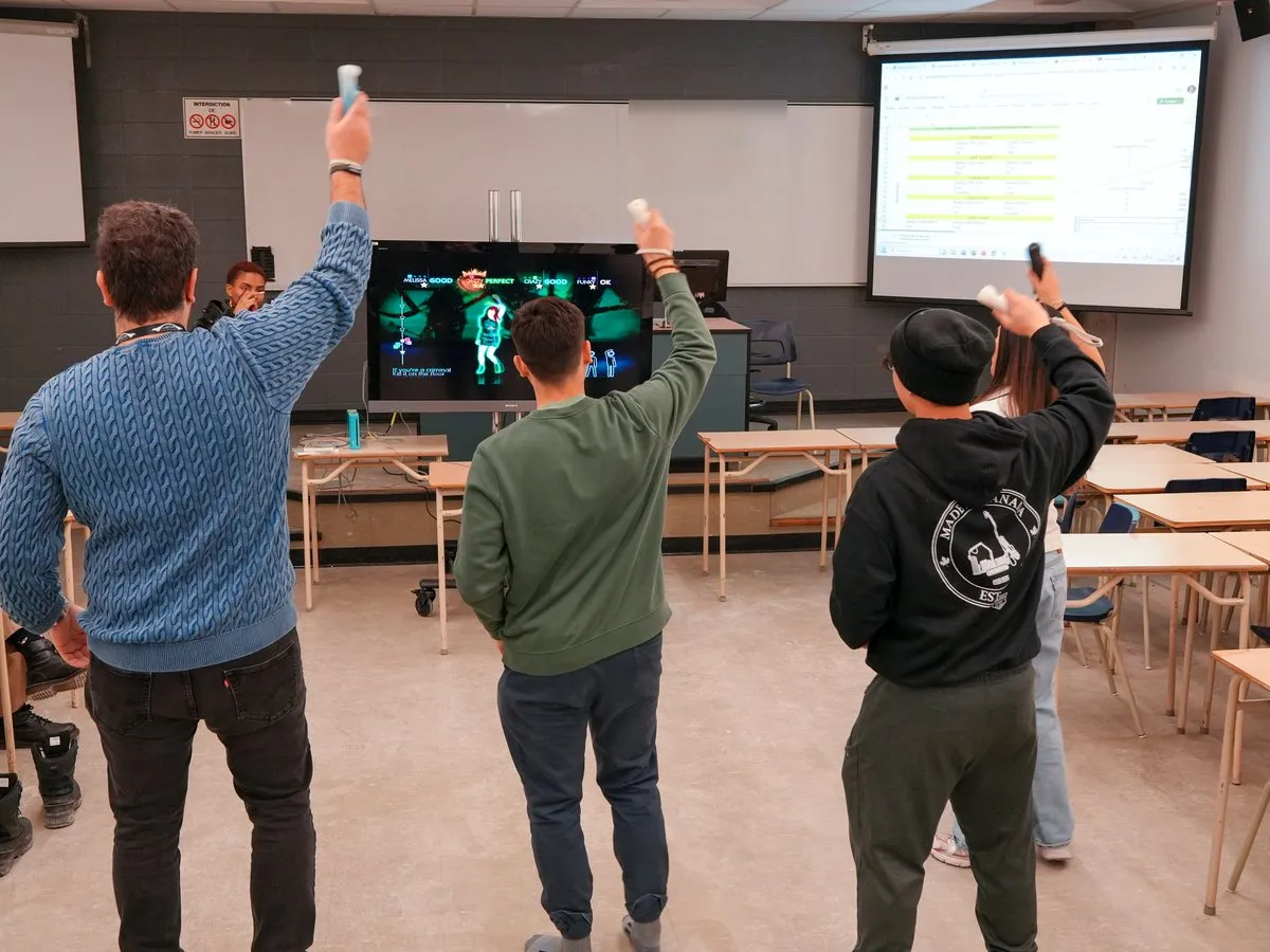 Four students raise game controllers as they dance in front of a TV showing Just Dance in a classroom.