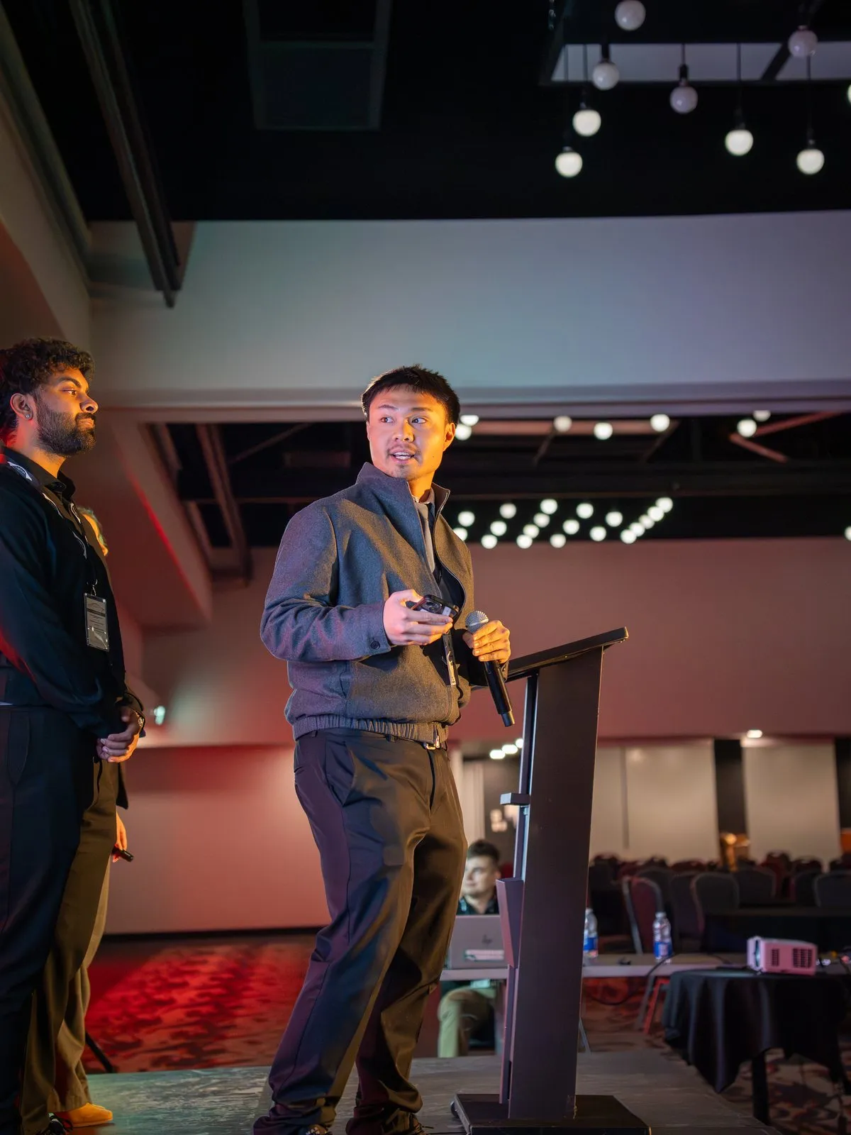 A student holding a microphone and slide clicker presents from a podium as a teammate looks on.